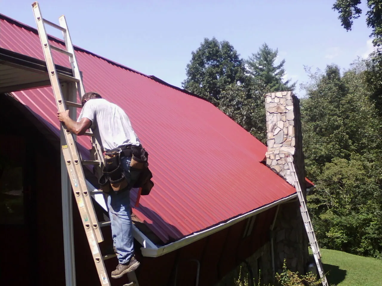 Roofer on a ladder beside a red metal roof and stone chimney — Soffit & Fascia Repair by Roofing Brothers in Pelion